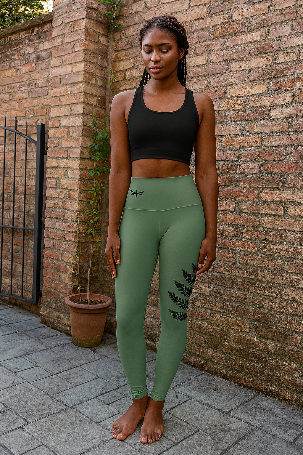 Woman wearing green leggings with a leaf design and black sports bra against a brick wall.