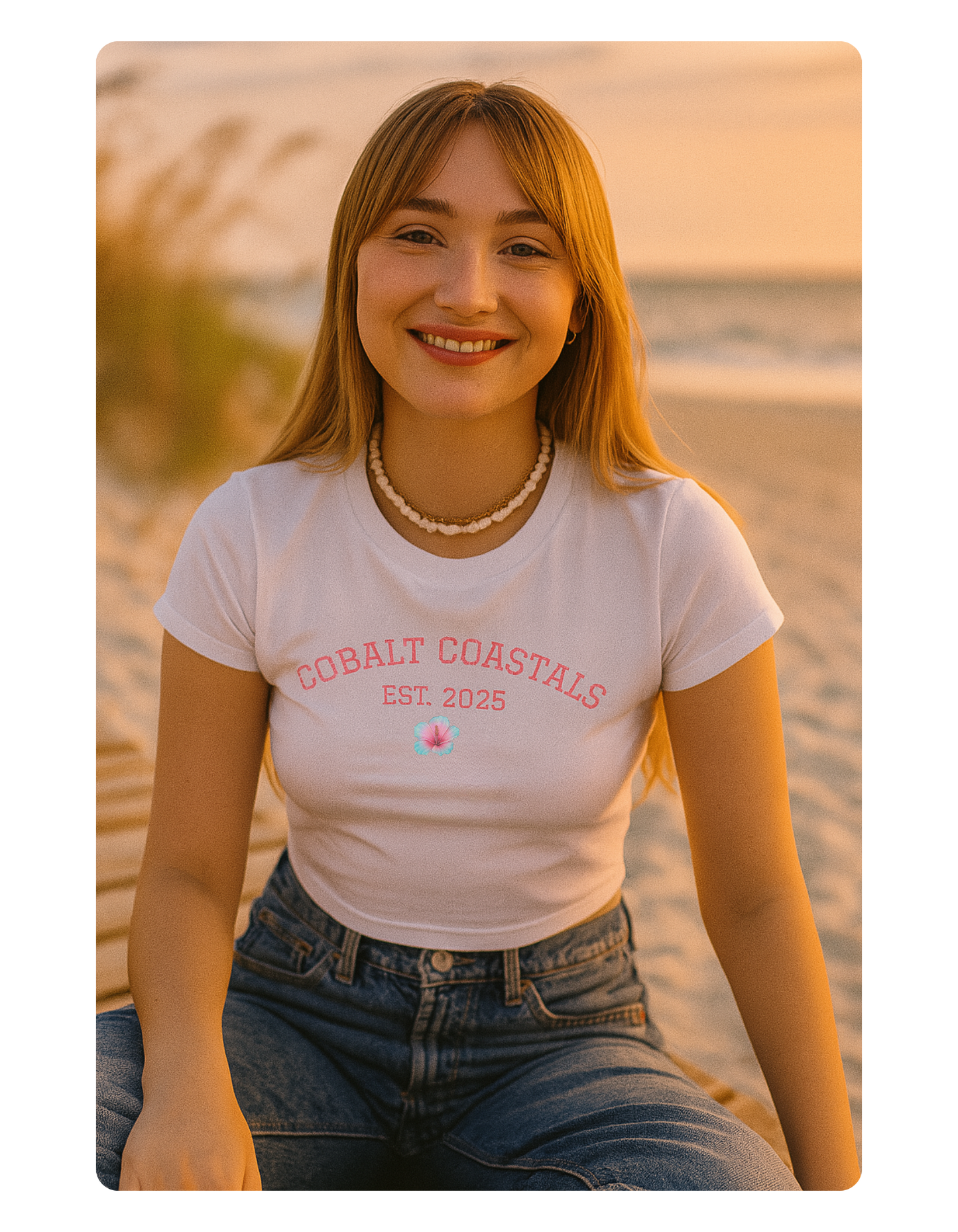 Woman wearing a white t-shirt with 'Coastal Coasts' text sitting on a beach at sunset.