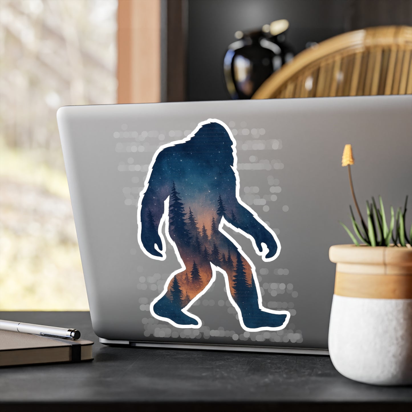 Laptop with a Bigfoot silhouette sticker on a desk with a plant and pen.