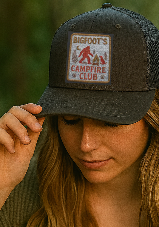Woman wearing a cap with a 'Bigfoot's Campfire Club' patch against a blurred natural background