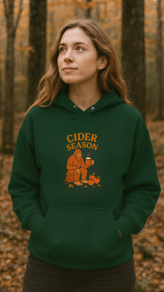 Person wearing a green hoodie with 'Cider Season' and big foot sasquatch design in a forest setting