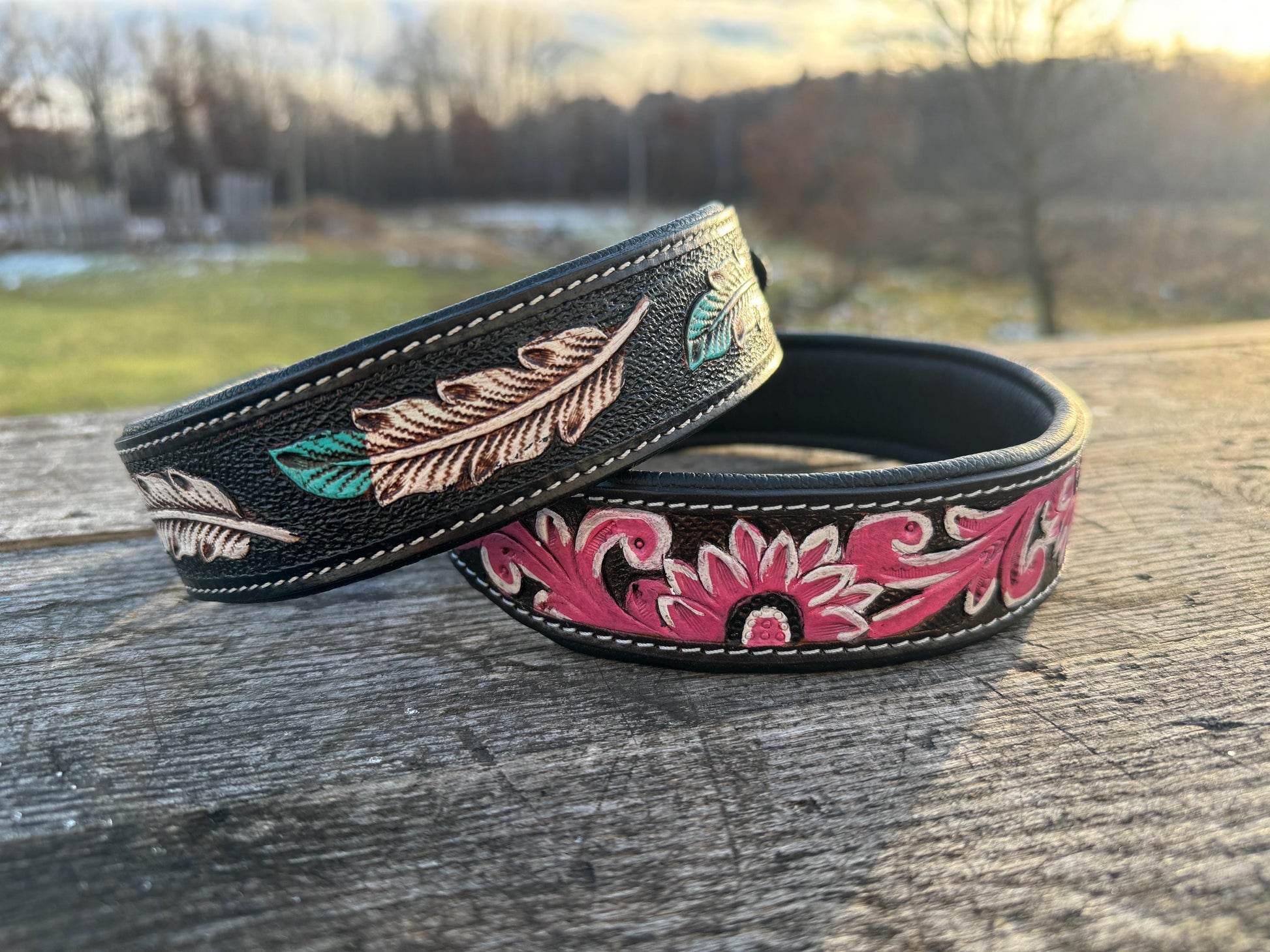 Two leather dog collars with floral and feather designs on a wooden surface.