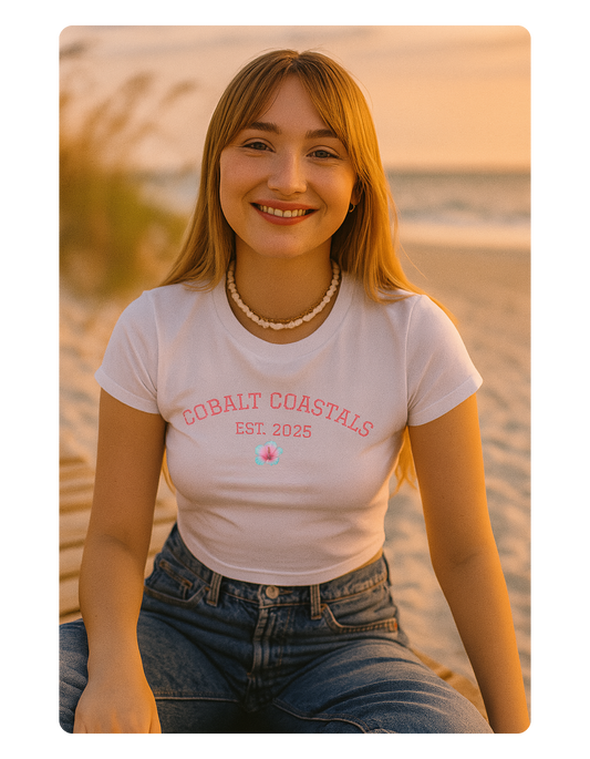 Woman wearing a white t-shirt with 'Coastal Coasts' text sitting on a beach at sunset.